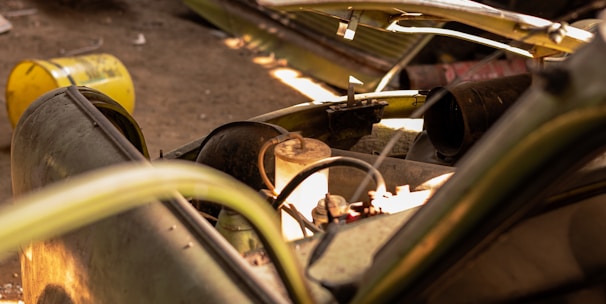 A dismantled and abandoned vehicle is situated in a dimly lit garage. The vehicle's hood is removed, exposing its internal components. Rusty barrels and a bench are in the background, along with other discarded items. Sunlight filters in, casting shadows across the dusty floor.